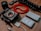 A collection of photography equipment is arranged on a wooden surface, including a black digital camera body without a lens, a red coiled cable, camera batteries, a flexible tripod, a camera lens, a USB cable, and LED light panels.