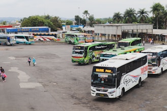 A busy bus terminal with multiple buses parked in queues. The buses are in various colors, including green, blue, and white, with logos and advertisements on their sides. There are a few people walking around, some carrying bags. In the background, there is a small building with a sign reading 'ISTIRAHAT SUPIR', surrounded by trees and greenery.