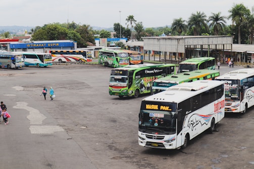 A busy bus terminal with multiple buses parked in queues. The buses are in various colors, including green, blue, and white, with logos and advertisements on their sides. There are a few people walking around, some carrying bags. In the background, there is a small building with a sign reading 'ISTIRAHAT SUPIR', surrounded by trees and greenery.