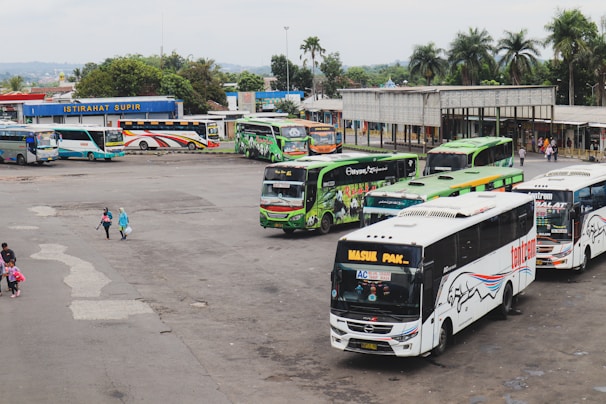 A busy bus terminal with multiple buses parked in queues. The buses are in various colors, including green, blue, and white, with logos and advertisements on their sides. There are a few people walking around, some carrying bags. In the background, there is a small building with a sign reading 'ISTIRAHAT SUPIR', surrounded by trees and greenery.
