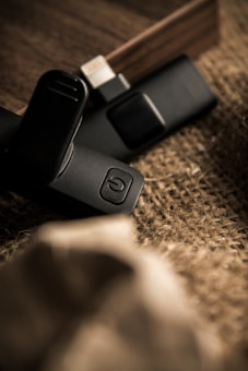 Two electronic devices rest on a textured burlap surface, with a warm wooden background. The focus is on a black device with a power button symbol, partially blurred in the foreground.