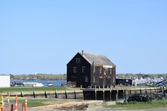 A historic wooden building with the sign 'Sail Loft' stands on a pier by the water. The structure is dark brown, with multiple windows and a pitched roof. Nearby, several orange traffic cones are visible, and beyond the building, a body of water and a tree-lined shore stretch into the distance.