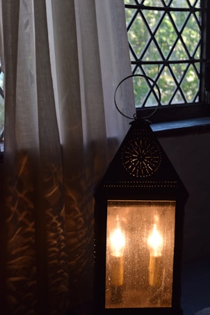 A dimly lit vintage-style lantern with two candles inside casts warm light. It is placed near a window with diamond-patterned panes, alongside a light-colored curtain that partially reflects the lantern's glow. The setting is calm and slightly rustic, with natural light filtering through the window.