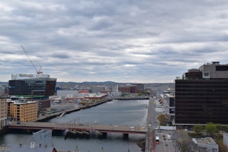 A cityscape featuring a river lined with modern buildings on either side. A construction crane is visible over one of the structures, indicating ongoing development. The sky is overcast with thick clouds, and a bridge crosses the river in the foreground. Roads and streets are visible, with a few vehicles and trees lining the area.