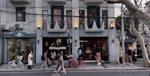 A stylish urban street scene with people walking past a row of modern shops. The façade is adorned with contemporary signage for 'LIU MING TANG' and 'INDIGO LIVING', and a large Coach logo is displayed in one window. A red bicycle is parked against the wall, and a small dog on a leash is walking with its owner. The building exterior is a cool gray, with architectural detailing and air conditioning units visible.