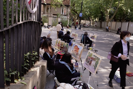 A group of people are sitting on the sidewalk engaged in painting, with easels set up displaying colorful artwork. The scene takes place on a tree-lined street with a few pedestrians passing by. The setting is calm with a mix of natural and urban elements.