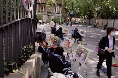 A group of people are sitting on the sidewalk engaged in painting, with easels set up displaying colorful artwork. The scene takes place on a tree-lined street with a few pedestrians passing by. The setting is calm with a mix of natural and urban elements.