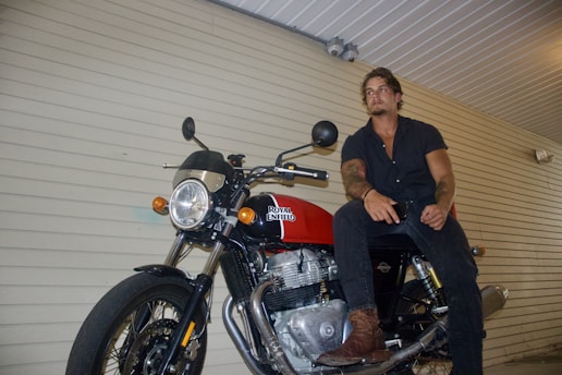A person is seated casually on a motorcycle in a covered area with a beige horizontal-panel wall. The motorcycle is a Royal Enfield with a prominent red fuel tank. The person is dressed in a dark shirt and pants, wearing brown boots. The setting appears like a simple parking or storage area.