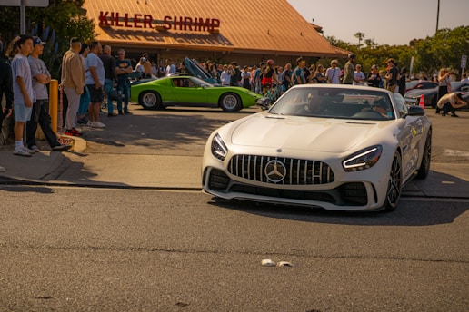 A white Mercedes convertible is prominently situated in the foreground, driving on a street. In the background, a group of people are gathered near a restaurant called Killer Shrimp. A green sports car with open doors is parked nearby, adding to the event's atmosphere.