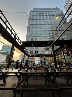 A rooftop outdoor dining area with wooden tables and benches is partially occupied by a group of people. Overhead, there are string lights, and in the background, a tall building and other urban structures are visible. The sky appears clear as the sun sets, casting a warm hue.