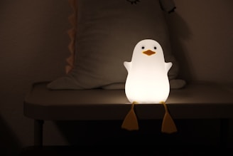 A small penguin-shaped night light sits on a table, emitting a warm glow. The penguin has a smooth white body with an orange beak and feet, and is positioned in a dimly lit room with a soft, minimalistic backdrop.