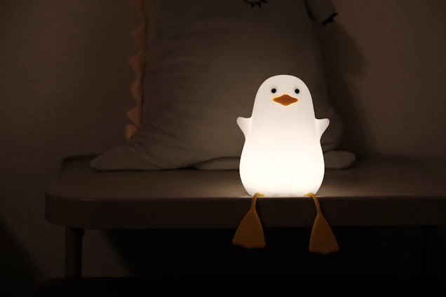A small penguin-shaped night light sits on a table, emitting a warm glow. The penguin has a smooth white body with an orange beak and feet, and is positioned in a dimly lit room with a soft, minimalistic backdrop.