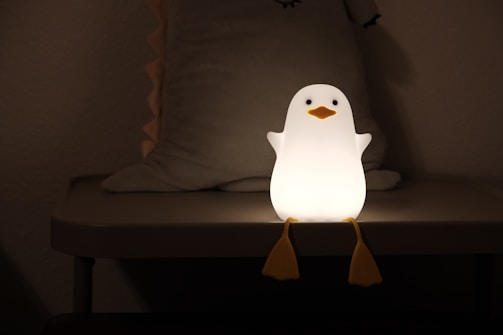 A small penguin-shaped night light sits on a table, emitting a warm glow. The penguin has a smooth white body with an orange beak and feet, and is positioned in a dimly lit room with a soft, minimalistic backdrop.