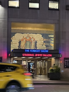 A theater entrance featuring a colorful mural above, with a prominent sign in neon lights reading 'The Tonight Show Starring Jimmy Fallon.' Below the sign, the entrance doors are visible, and there is a person walking by. A yellow taxi is in motion in the foreground, adding a dynamic element to the scene.