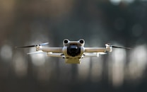 A drone is flying in the air with its rotors clearly visible. It appears to be metallic with a sleek, modern design. The background is blurred, suggesting the photo was taken with a wide aperture, emphasizing the drone as the focal point.