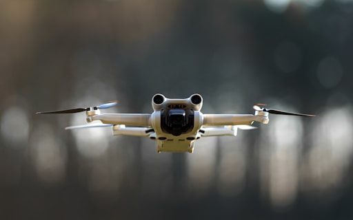 A drone is flying in the air with its rotors clearly visible. It appears to be metallic with a sleek, modern design. The background is blurred, suggesting the photo was taken with a wide aperture, emphasizing the drone as the focal point.