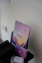A colorful calendar standing on a desk with a design that features abstract wave patterns and the words 'Joyful Obedience'. Below the calendar is a smaller card with the text 'Overwhelming Love'. Nearby, there is a black electronic device resembling a printer or scanner, and several sheets of paper are placed on the desk.