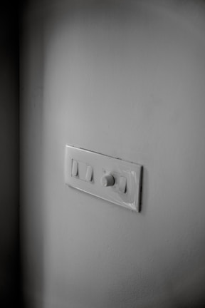 A white wall features a light switch panel with two switches and a rotary dimmer. The panel is positioned slightly off-center, and the lighting casts soft shadows around it. The surface of the wall and panel is clean and smooth.