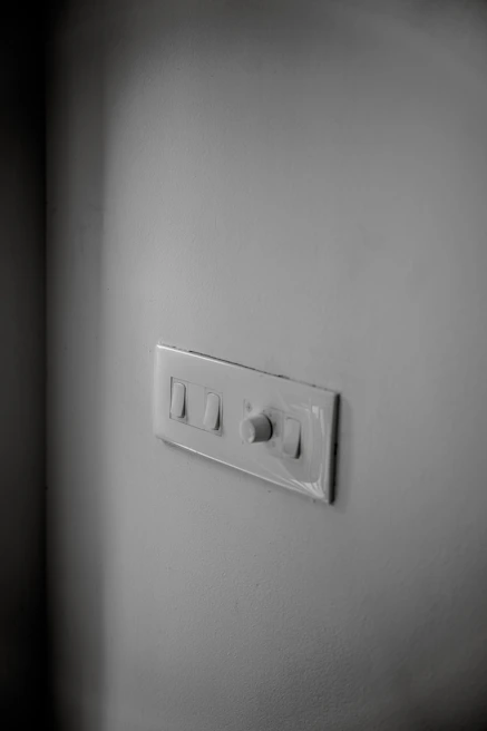 A white wall features a light switch panel with two switches and a rotary dimmer. The panel is positioned slightly off-center, and the lighting casts soft shadows around it. The surface of the wall and panel is clean and smooth.