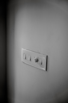 A white wall features a light switch panel with two switches and a rotary dimmer. The panel is positioned slightly off-center, and the lighting casts soft shadows around it. The surface of the wall and panel is clean and smooth.