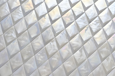 A pattern of iridescent, diamond-shaped tiles creating a textured surface with reflective qualities. The tiles are aligned in a grid with subtle color variations and swirling patterns visible on their surfaces.