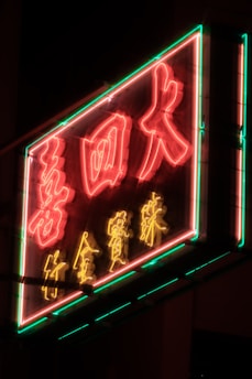 A vibrant neon sign featuring Chinese characters illuminated in bright red and yellow colors with an outline of green neon lights. The sign is slightly blurry, enhancing the glowing effect.