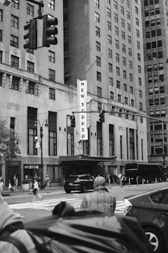 A busy urban street scene features a tall building with a large vertical sign reading 'NEW YORKER' and 'WYNDHAM'. Pedestrians cross the street, and several vehicles including cars and a bus are visible. A traffic light hangs in the foreground, and the building's architecture has a classic design with rectangular windows and decorative elements.