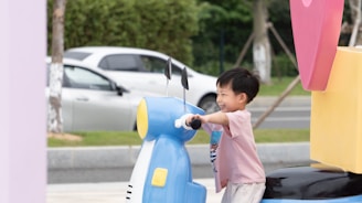 A young child is joyfully playing on a colorful, oversized toy vehicle. He is smiling broadly, gripping the handles and appearing to be in motion. In the background, a white car is parked near a roadside with greenery.
