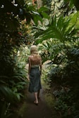 A person with blonde hair walks barefoot along a narrow, lush path surrounded by dense green foliage. The scene is tranquil and nature-focused, with various plants and leaves arching overhead and creating a canopy. The light filtering through the leaves adds a serene and natural ambiance.