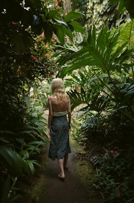 A person with blonde hair walks barefoot along a narrow, lush path surrounded by dense green foliage. The scene is tranquil and nature-focused, with various plants and leaves arching overhead and creating a canopy. The light filtering through the leaves adds a serene and natural ambiance.