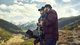 A film crew setting up cameras in a scenic valley surrounded by mountains.