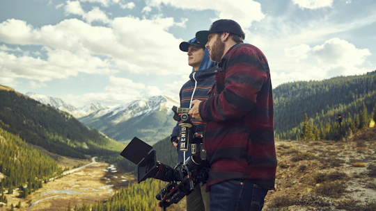 A film crew setting up cameras in a scenic valley surrounded by mountains.