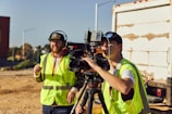 Two people wearing high-visibility vests are operating a professional video camera mounted on a tripod. They appear to be filming outdoors on a sunny day, with one person adjusting the camera and the other possibly giving directions. A large truck is visible in the background.