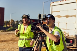 Two people wearing high-visibility vests are operating a professional video camera mounted on a tripod. They appear to be filming outdoors on a sunny day, with one person adjusting the camera and the other possibly giving directions. A large truck is visible in the background.