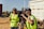 Two people wearing high-visibility vests are operating a professional video camera mounted on a tripod. They appear to be filming outdoors on a sunny day, with one person adjusting the camera and the other possibly giving directions. A large truck is visible in the background.