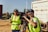 Two people wearing high-visibility vests are operating a professional video camera mounted on a tripod. They appear to be filming outdoors on a sunny day, with one person adjusting the camera and the other possibly giving directions. A large truck is visible in the background.