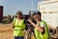 Two people wearing high-visibility vests are operating a professional video camera mounted on a tripod. They appear to be filming outdoors on a sunny day, with one person adjusting the camera and the other possibly giving directions. A large truck is visible in the background.