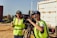Two people wearing high-visibility vests are operating a professional video camera mounted on a tripod. They appear to be filming outdoors on a sunny day, with one person adjusting the camera and the other possibly giving directions. A large truck is visible in the background.