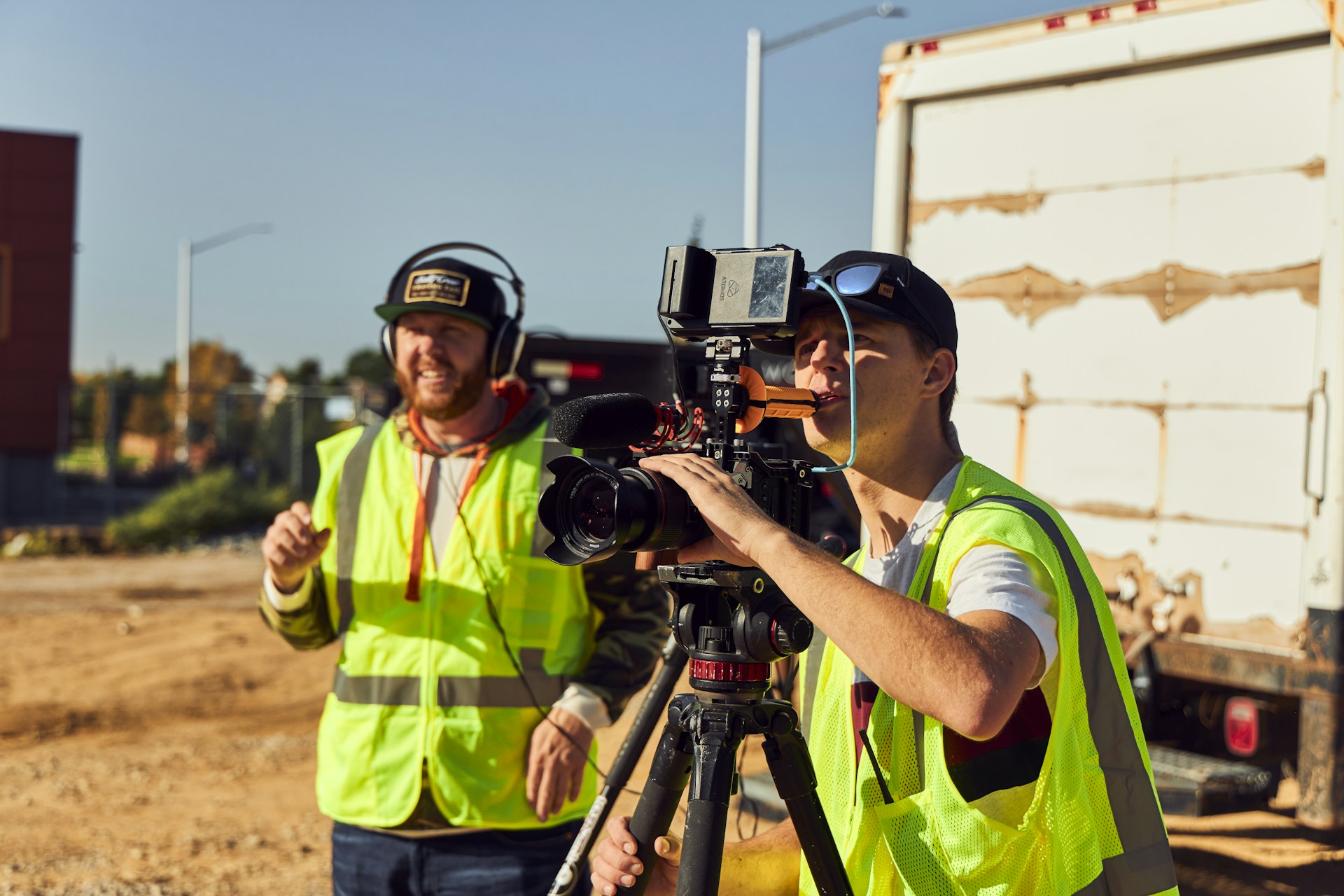 Two people wearing high-visibility vests are operating a professional video camera mounted on a tripod. They appear to be filming outdoors on a sunny day, with one person adjusting the camera and the other possibly giving directions. A large truck is visible in the background.