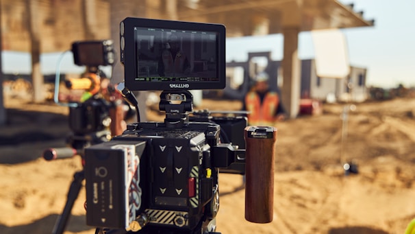 A professional video camera setup is positioned on a construction site. The camera has a small monitor attached, displaying a preview of the scene. In the background, there is a blurred figure wearing a safety vest, with construction equipment and a structure visible under a clear sky.