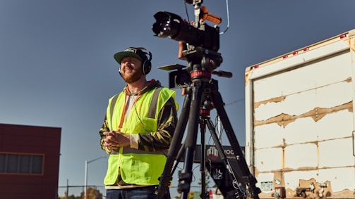 A person wearing a green safety vest and headphones stands outdoors, holding a communication device. A professional video camera is mounted on a tripod next to the person. The background includes an industrial area with buildings and a weathered vehicle.