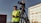 A person wearing a green safety vest and headphones stands outdoors, holding a communication device. A professional video camera is mounted on a tripod next to the person. The background includes an industrial area with buildings and a weathered vehicle.