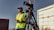 A person wearing a green safety vest and headphones stands outdoors, holding a communication device. A professional video camera is mounted on a tripod next to the person. The background includes an industrial area with buildings and a weathered vehicle.