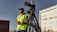 A person wearing a green safety vest and headphones stands outdoors, holding a communication device. A professional video camera is mounted on a tripod next to the person. The background includes an industrial area with buildings and a weathered vehicle.