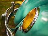 A close-up view of a vintage car's headlight, featuring a yellow lens surrounded by shiny chrome. The light blue car body with smooth curves is visible in the background, along with a reflective chrome bumper.