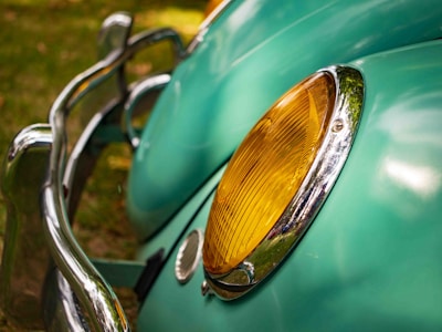 A close-up view of a vintage car's headlight, featuring a yellow lens surrounded by shiny chrome. The light blue car body with smooth curves is visible in the background, along with a reflective chrome bumper.