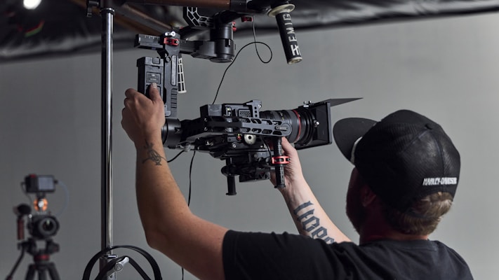 A camera operator is adjusting professional camera equipment, mounted on a rig. The person wears a black cap and has tattoos on their arm. The setup includes multiple components such as lenses and microphones, indicating a focus on video or film production.