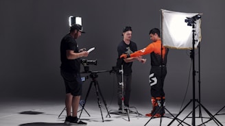 In a studio setting, three individuals are engaged in a photoshoot. One person is adjusting the attire of another, who is wearing a bright orange and black outfit, possibly for a motorsport or similar activity. The third person is checking a monitor or camera setup. The scene is illuminated by professional photography lighting equipment.