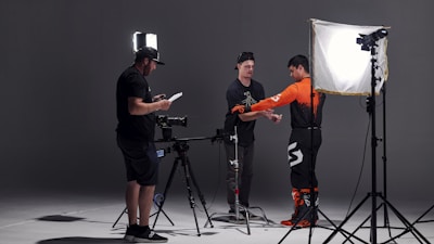 In a studio setting, three individuals are engaged in a photoshoot. One person is adjusting the attire of another, who is wearing a bright orange and black outfit, possibly for a motorsport or similar activity. The third person is checking a monitor or camera setup. The scene is illuminated by professional photography lighting equipment.
