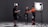 In a studio setting, three individuals are engaged in a photoshoot. One person is adjusting the attire of another, who is wearing a bright orange and black outfit, possibly for a motorsport or similar activity. The third person is checking a monitor or camera setup. The scene is illuminated by professional photography lighting equipment.